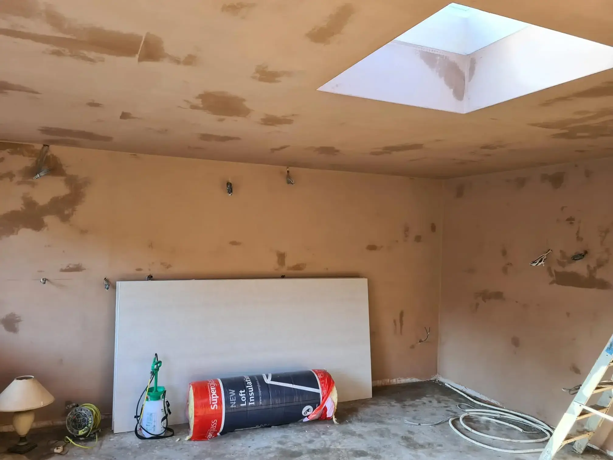 Room with walls and ceiling patched and prepared around a roof light, with plasterboard and insulation stacked ready for use.