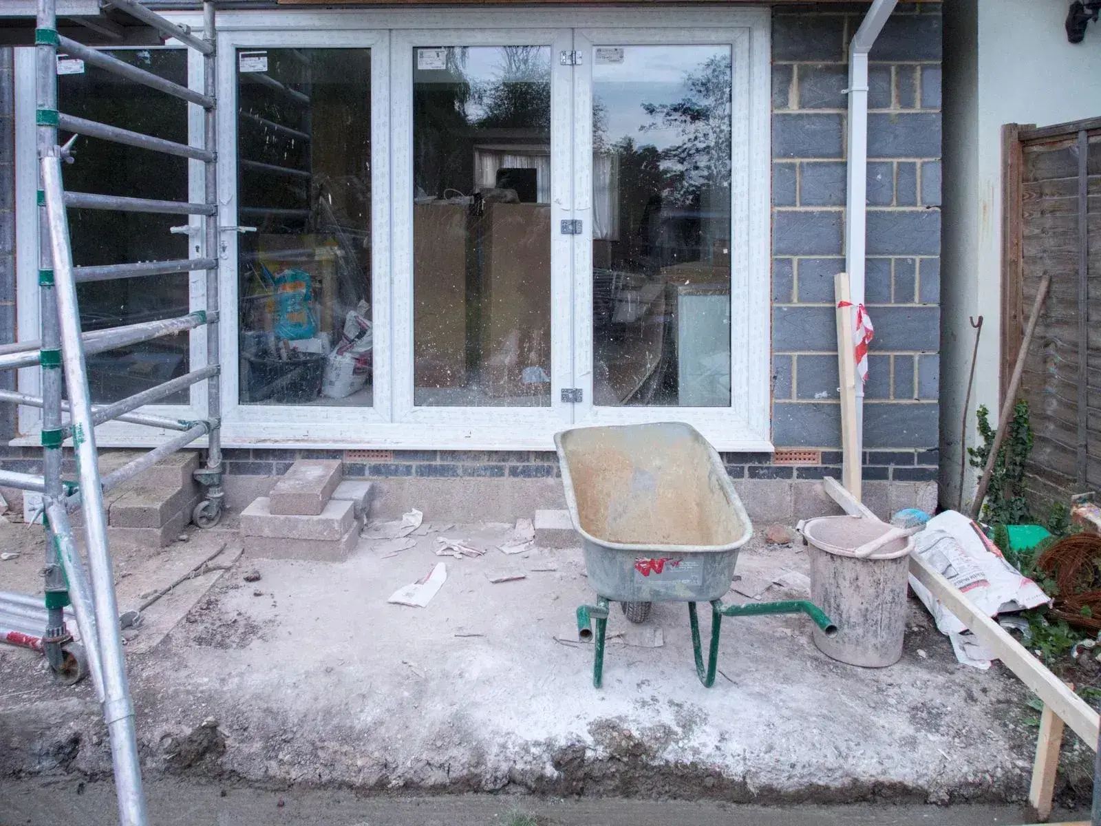 The back of a house part-way through building work, with new glazed doors fitted, blockwork exposed, and a scaffold tower, wheelbarrow and mixing tub standing on bare ground.
