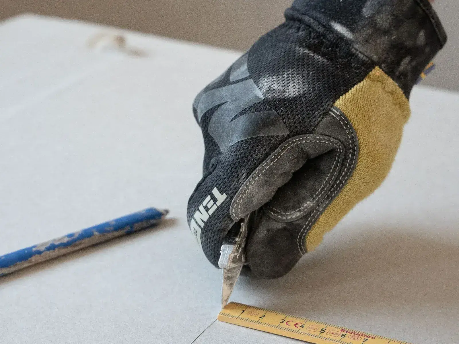 A gloved hand scoring a sheet of plasterboard with a knife, run along the edge of a folding rule.