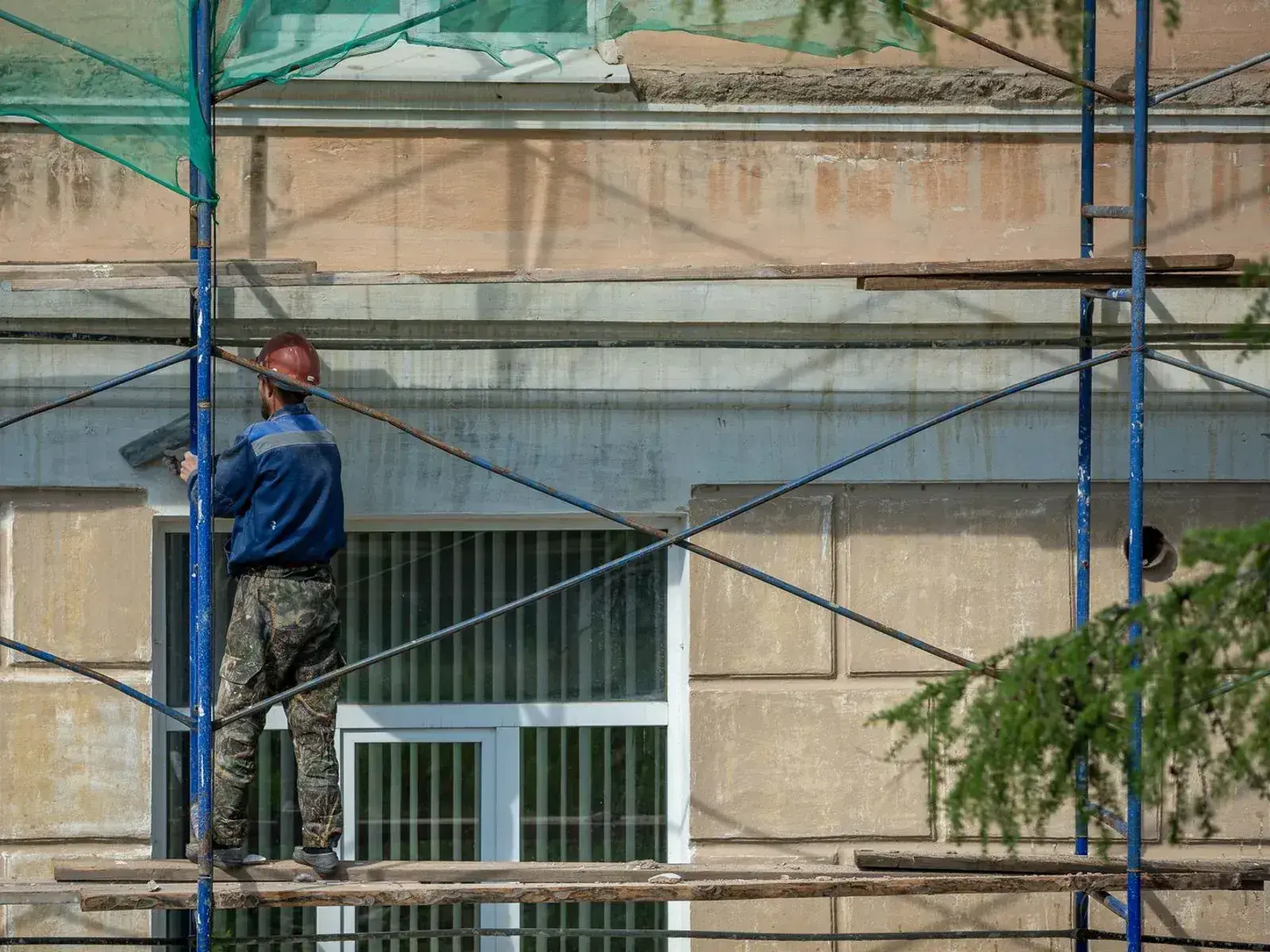 A worker standing on scaffolding, floating render onto the outside wall of a building above a window.